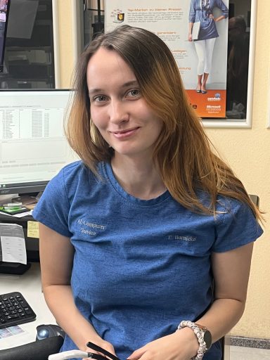 Junge Frau mit langen, braunen Haaren, lächelnd, in blauer Kleidung an einem Schreibtisch.